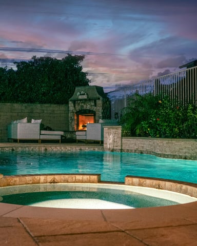 Glowing freeform pool with attached spa, stone fireplace, and dramatic purple sunset sky