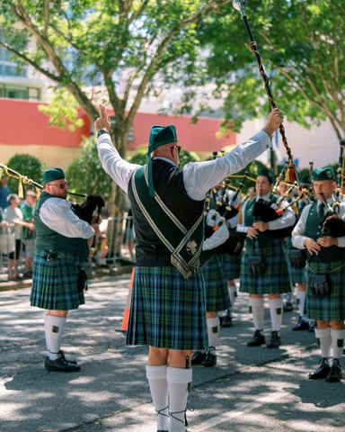 Bagpipe marching band in kilts parading down a sunny street under green trees
