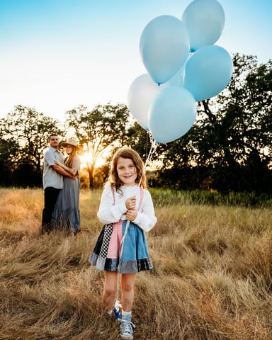 Family in a golden field at sunset, young girl holding blue balloons