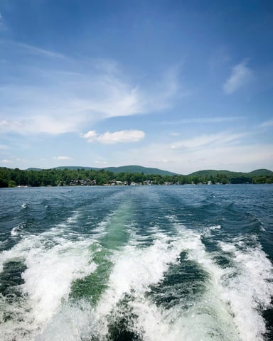 Boat wake view looking back across a green lake with wooded hills and blue sky