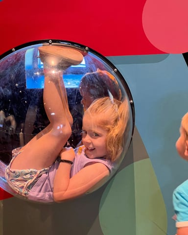 Girl laughing inside a bubble porthole at a children's museum with colorful red and blue walls