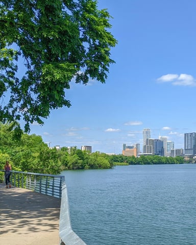 Lady Bird Lake trail with a person on the walkway, lush trees, and the Austin downtown skyline across the water