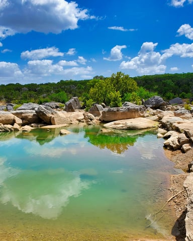 Turquoise Hill Country swimming hole with limestone boulders and lush green trees under a blue sky