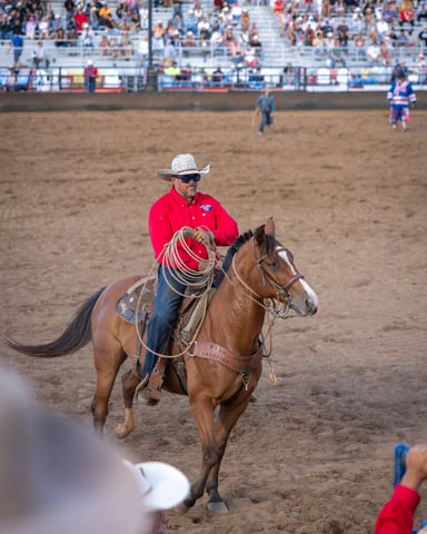 Cowboy in red shirt on horseback with lasso, packed grandstands behind him at a rodeo arena