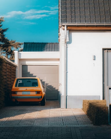 Orange car parked on a cobblestone driveway beside a white home with a garage door and blue sky