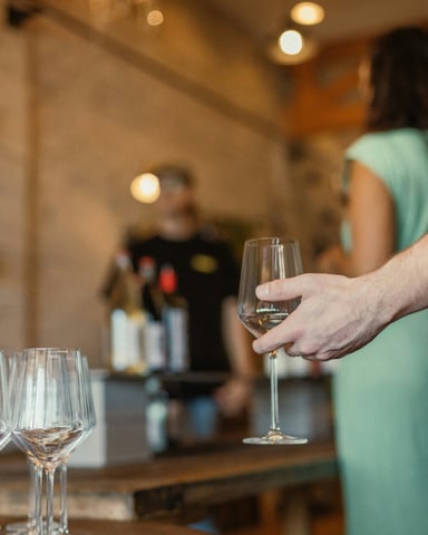 Hand holding a wine glass at a tasting event, pourer and bottles visible behind, warm pendant lights