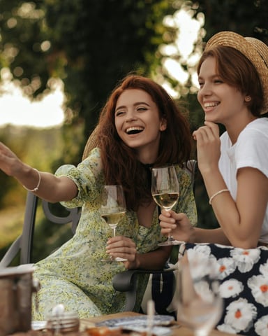 Two women laughing and clinking white wine glasses outdoors in a garden