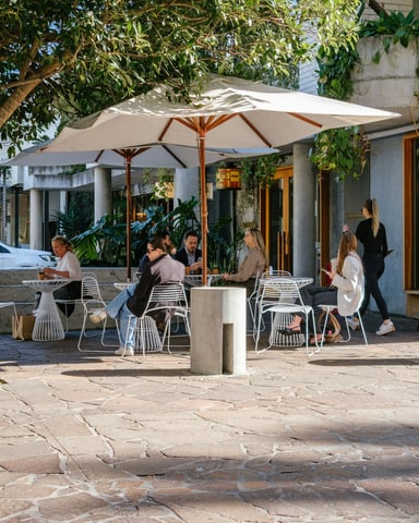Bright sunny outdoor cafe patio with white umbrellas, people dining, and lush trees