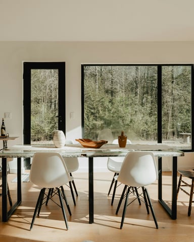 Bright vacation home dining room with long table and floor-to-ceiling windows looking out to trees