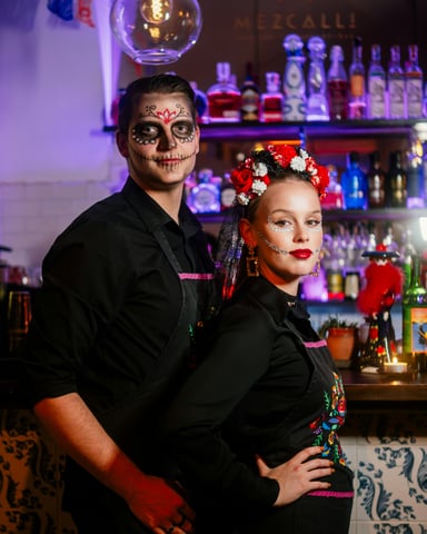 Couple in Day of the Dead face paint and costumes posing at a bar with bottles behind them