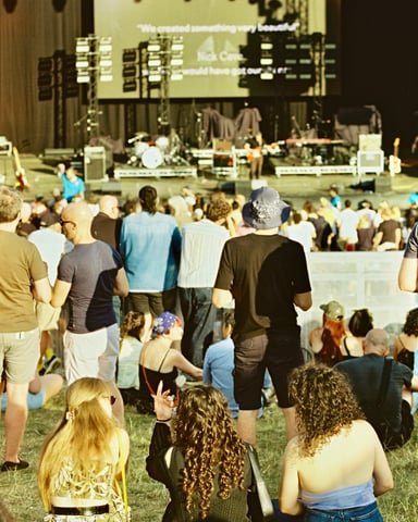 Crowd sitting on grass watching an outdoor music festival stage at dusk