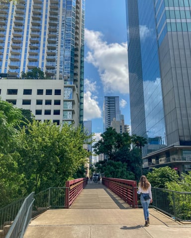 Woman walking across a pedestrian bridge over Shoal Creek with Austin downtown towers rising behind her