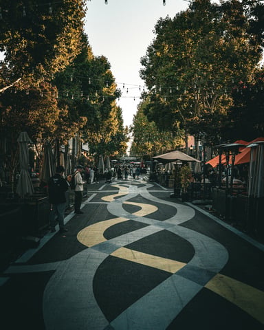 Tree-lined outdoor shopping promenade with string lights, outdoor dining umbrellas, and patterned pavement