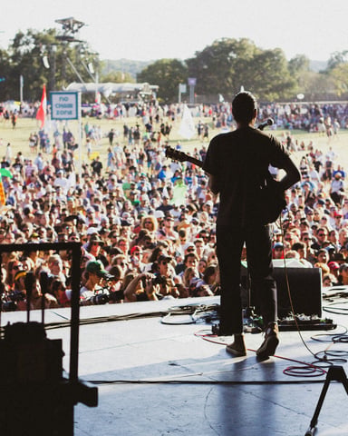 Performer on stage facing a massive outdoor crowd on a sunny Texas lawn at ACL