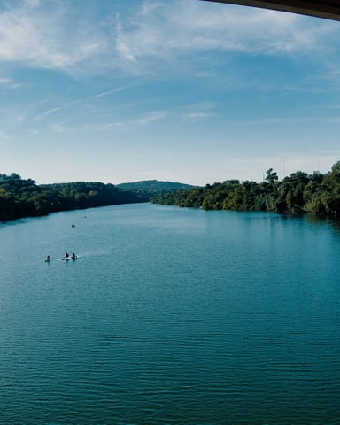 Calm lake with paddleboarders under a wide blue sky and lush green tree-lined shores