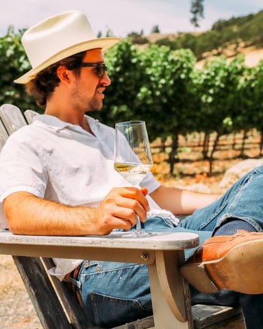 Man in a straw hat relaxing in an Adirondack chair with a glass of white wine, vineyard rows behind him