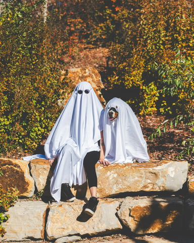 Two people in ghost costumes sitting on a sunny rock wall surrounded by fall foliage
