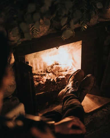 Person relaxing with boots up on a fireplace hearth, warm amber glow from the fire