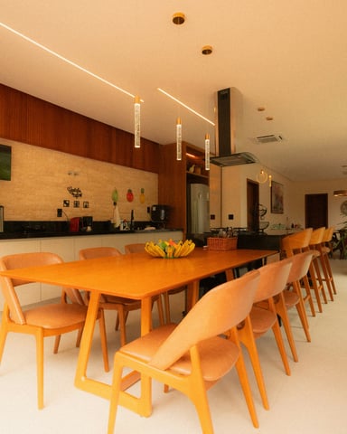 Long dining table seating 10+ guests in a modern open-plan home with floor-to-ceiling glass walls and lush greenery outside