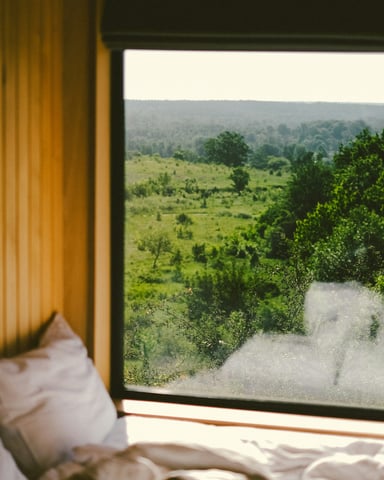 Bed with white pillows framed by a wide window overlooking a lush green Hill Country landscape