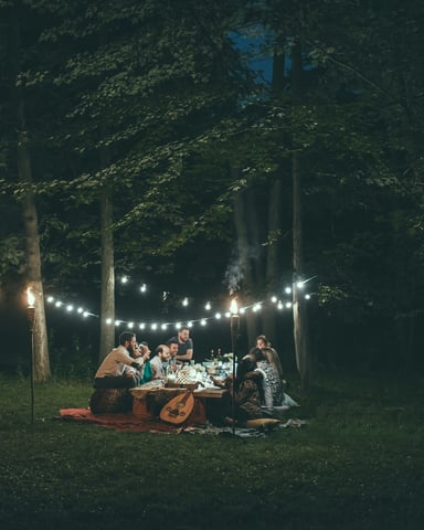 Group gathered around a long outdoor dinner table under string lights in a wooded setting at night