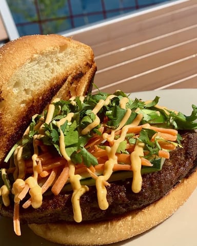 Gourmet burger with beef patty, coleslaw, and sauce on toasted bun, served on wooden deck table
