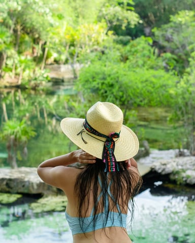 Woman in straw hat overlooking lush jungle cenote with clear water and dense green vegetation