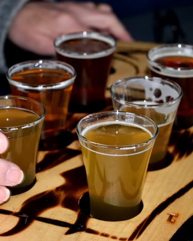 Beer tasting flight with six glasses of varying amber and dark beers on wooden paddle, person holding one glass
