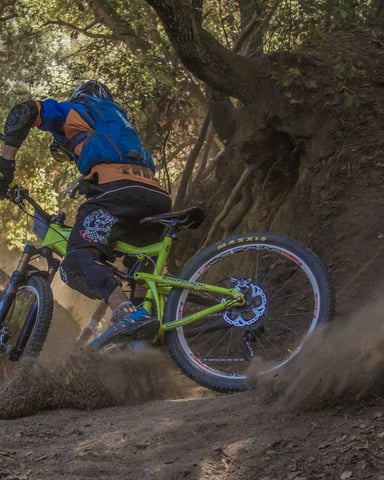 Mountain biker in blue helmet and gear riding lime-green bike on dusty trail through tree-lined canyon.