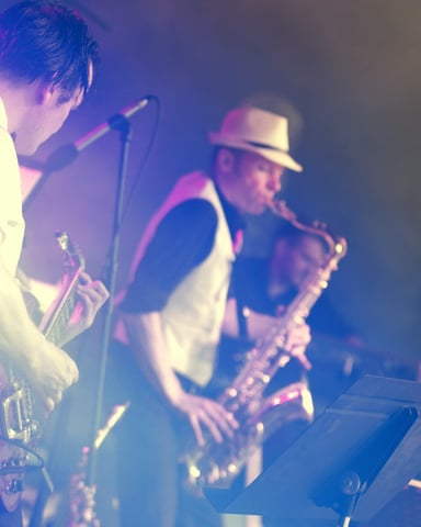 Live band performing on stage with blue and pink lighting, guitarist in foreground and saxophonist wearing hat