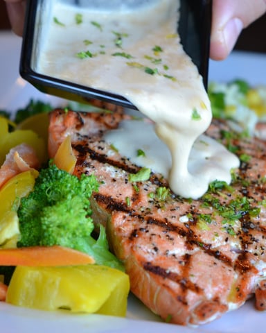 Grilled salmon steak with creamy sauce being drizzled on top, served with broccoli and diced vegetables