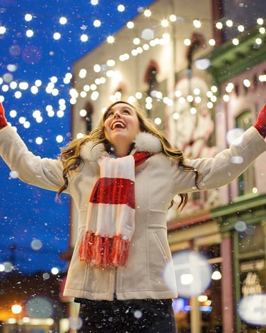 Woman in white coat and red mittens with arms raised in falling snow on decorated holiday street at night