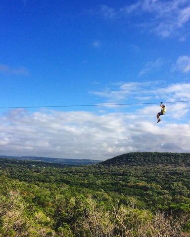 Person zip-lining across forested valley with blue sky and white clouds