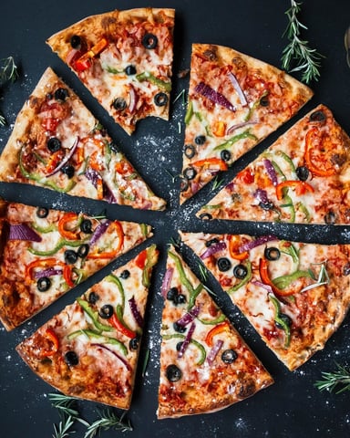 Sliced pizza with cheese, ham, olives, and vegetables on dark surface with fresh ingredients and beer nearby