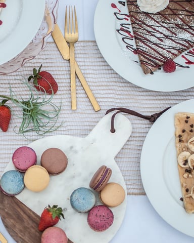 Overhead view of dessert plates with crepes, chocolate cake, macarons, and fruit on striped tablecloth with gold utensils