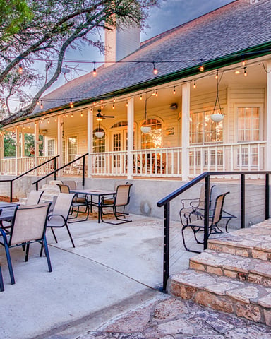 Stone house with wraparound porch, outdoor patio seating, string lights, and blooming tree at dusk