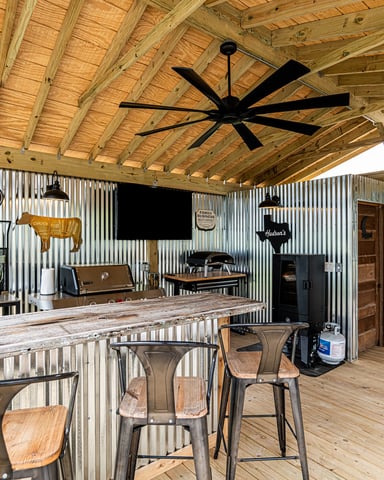 Rustic covered patio with wood beams, corrugated metal walls, bar counter, black ceiling fan, and vintage signage