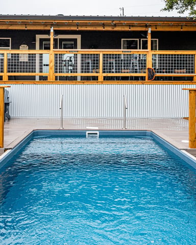 Rectangular swimming pool with wooden deck and metal railing, modern black house with stone chimney in background