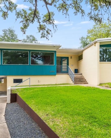 Mid-century modern house with turquoise and cream exterior, large windows, carport, and manicured lawn surrounded by trees