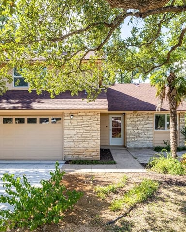 Stone single-story house with two-car garage, shaded by large mature trees with green foliage