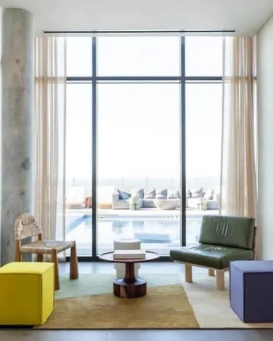 Modern living room with floor-to-ceiling windows overlooking rooftop pool, colorful seating, and wall-mounted TV