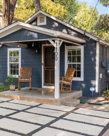 Modern dark blue cottage with covered porch, white trim, rocking chairs, and concrete paver pathway through landscaped yard with tall trees.