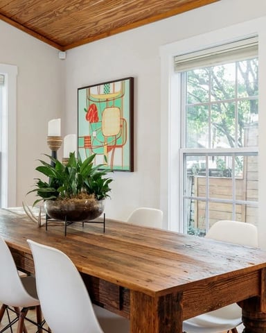 Dining room with wooden table, white chairs, potted plant, and large windows overlooking trees