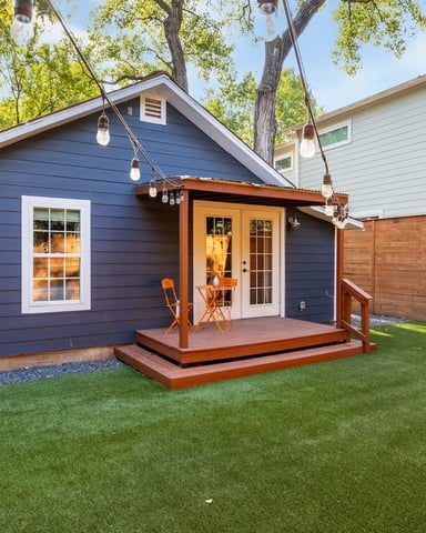 Modern blue cottage with wooden deck, glass doors, and hanging lights surrounded by mature trees and manicured lawn