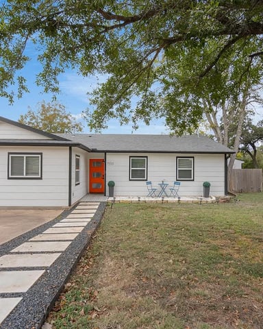 Single-story white house with red door, driveway, and large shaded yard with mature trees