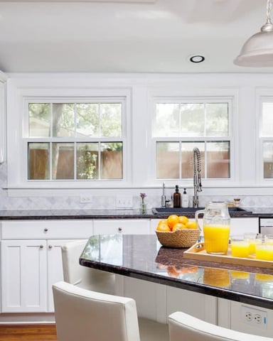 Bright white kitchen with black granite countertops, stainless steel refrigerator, island with seating, and multiple windows overlooking trees.