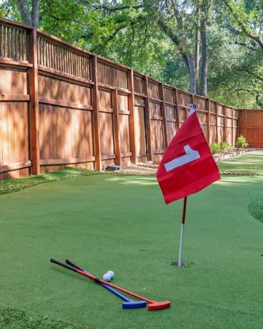Backyard mini golf course with red flag, putter, and ball on green turf, wooden fence and tall trees