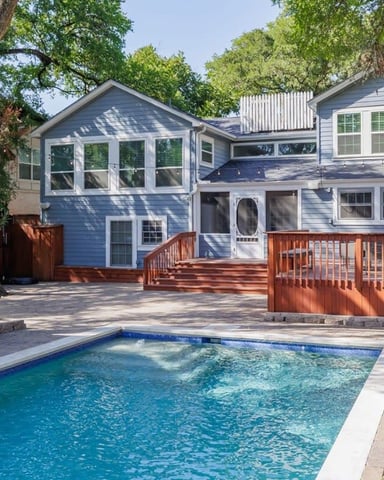 Blue two-story house with wooden deck and swimming pool surrounded by mature trees