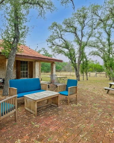 Stone house with patio seating area featuring wooden furniture with blue cushions under large oak trees