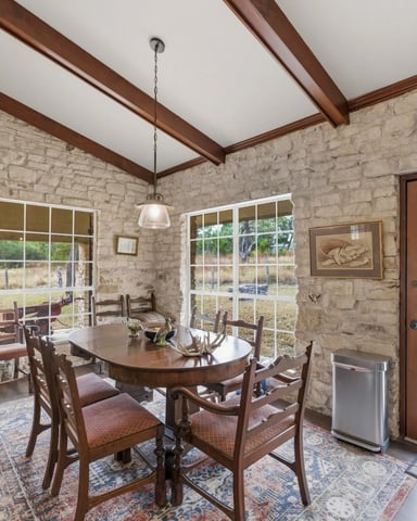 Dining room with stone walls, exposed wood beams, round wooden table, ladder-back chairs, and glass doors opening to pasture